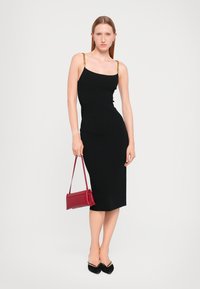 Black fitted dress with thin spaghetti straps featuring gold chain accents. Model holds a small red handbag and wears black pointed flats.