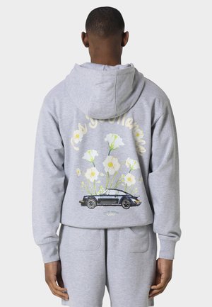 Uomo che indossa una felpa con cappuccio grigio chiaro e pantaloni della tuta, sul retro una grafica di un'auto d'epoca nera con fiori bianchi e testo in corsivo sopra.