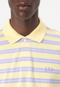 Polo shirt jaune avec des rayures verticales violettes et bleu clair, dotée d'un col, de deux boutons et d'un logo "GAP" brodé en violet.