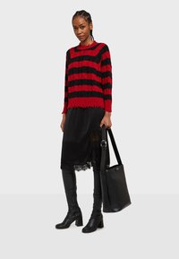 Červený a čierny pruhovaný pletený sveter s rozstrapkanými okrajmi, skombinovaný s čiernou sukňou s čipkovými detailmi a čiernymi koženými čižmami po kolená na vysokom podpätku.