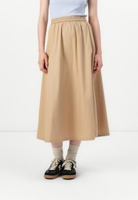 Falda beige hasta los tobillos con una cintura elástica, hecha de un tejido suave. Llevada con zapatillas negras y calcetines de color crema.