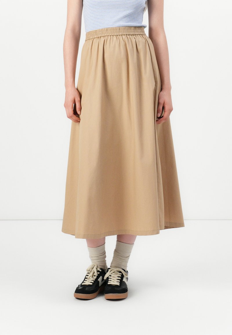 Falda beige hasta los tobillos con una cintura elástica, hecha de un tejido suave. Llevada con zapatillas negras y calcetines de color crema.