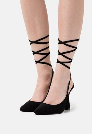 Scarpe nere in suede con tacco alto, punta affusolata, apertura sul retro e fasce incrociate alla caviglia. Presentano un tacco robusto per garantire stabilità.