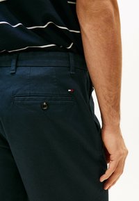 Pantaloni navy con texture, dotati di una tasca posteriore con bottone e un piccolo accento di logo rosso, bianco e blu. Passanti per la cintura visibili.