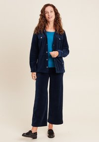 Marineblaue Cordjacke mit vorderen Taschen, kombiniert mit einer passenden Hose im weiten Schnitt und einem türkisfarbenen Top. Schwarze Slipper runden das Outfit ab.