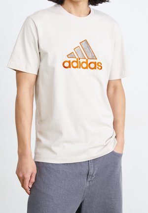 Person wearing a beige Adidas T-shirt with orange logo, paired with gray pants, one hand in pocket and the other relaxed by the side.