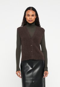 Brązowa, puszysta kamizelka z dekoltem w serek, z czterema brązowymi guzikami, noszona na długim, ciemnozielonym golfie. Poniżej czarna skórzana spódnica.