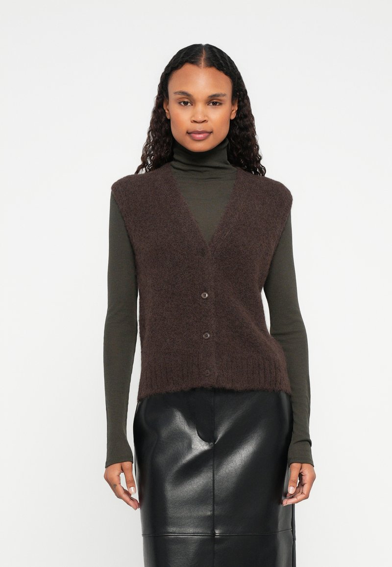 Brązowa, puszysta kamizelka z dekoltem w serek, z czterema brązowymi guzikami, noszona na długim, ciemnozielonym golfie. Poniżej czarna skórzana spódnica.