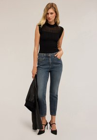 Top senza maniche a coste nere con pannello trasparente, abbinato a jeans high-waisted azzurri chiari e tacchi a stiletto neri. Tiene in mano una giacca di pelle nera.