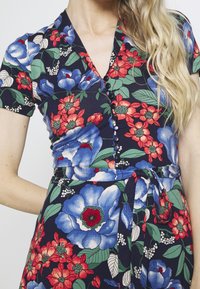 Donna che indossa un vestito blu navy con ampi motivi floreali blu e rossi, maniche corte, bottoni frontali e una cintura annodata in vita.