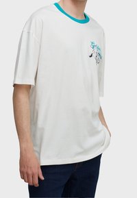 Hombre vestido con una camiseta blanca de manga corta con cuello turquesa y un pequeño gráfico con texto en el lado izquierdo del pecho, combinado con jeans oscuros.