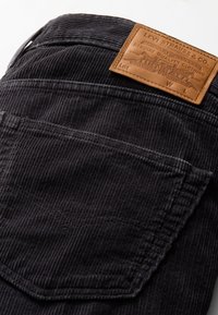 Zwarte corduroy stof met textuurlijke verticale strepen, voorzien van een tan leren label met branding op de tailleband en een detail van een achterzak.