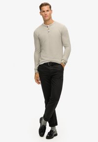 Lichtgrijs, langwerpig henley-shirt met drie knopen, gecombineerd met zwarte broek en zwarte schoenen, met grijze sokken.