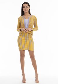 Keltainen ja violetti raidallinen neuletakki vaalean violetin paidan päällä, yhdistettynä samansävyiseen kapeaan hameeseen. Sileä pinta, slim fit -muotoilu, hienovarainen peplum-yksityiskohta.