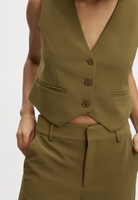 Nahaufnahme einer Person, die eine olivgrüne ärmellose Weste mit Knöpfen und passende hochgeschnittene Hose trägt, während sie die Hände in den Taschen hat, vor einem weißen Hintergrund.