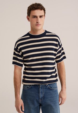 Młody mężczyzna stoi, ubrany w granatowy, biały i beżowy pasiasty krótki sweter oraz niebieskie dżinsy na jednolitym tle.