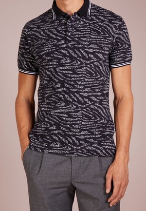 Mann trägt ein kurzärmeliges Polo-Shirt mit schwarz-weißem Muster und gestreiftem Kragen und Bündchen, kombiniert mit grauen Hosen, Hände in den Taschen.