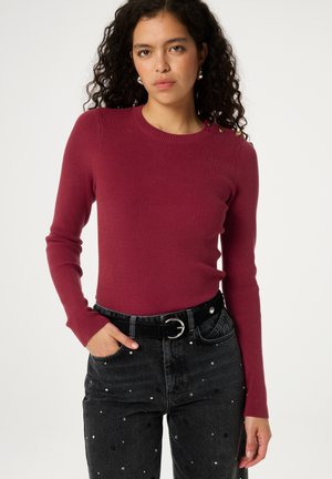 Bordeaux ribgebreide langwerpige top met een ronde halslijn, gecombineerd met zwarte jeans met decoratieve pailletjes en een zwarte riem met een zilveren gesp.