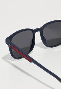 Ochelari de soare bleumarin cu lentile gri închis, un cadru gros și un accent roșu pe braț. Designul este unghiular, cu o finisare mată.