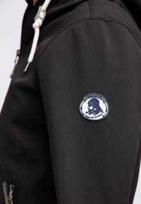 Svart softshell-jacka med rund, blå broderad lapp på ärmen med en karaktärsdesign, vita dragkedjor och fickor.