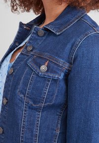 Donkerblauwe denimjas met een kraag, twee borstzakken, knoopsluitingen en zichtbare stikdetails. Glad textuur en een aansluitend ontwerp.