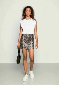 Top branco sem mangas, saia mini com padrão de cobra metálico e tênis brancos. Bolsa preta na mão, em pé sobre um fundo simples.