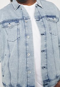 Vaaleansininen denim-takki, jonka muotoilu on rento. Takissa on kaksi rintataskua, napit ja näyttävät ompeluonnit.