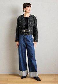 Veste cropped noire texturée avec des boutons argentés, associée à un jean en denim à jambes larges avec des poignets gris clair et des bottes pointues noires.