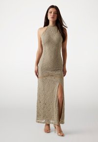 Tight-fitting beige lace evening gown with a high collar, open back, side slit, and intricate leaf-patterned texture. Stiletto heels complement the look.