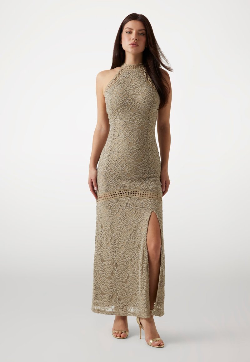 Tight-fitting beige lace evening gown with a high collar, open back, side slit, and intricate leaf-patterned texture. Stiletto heels complement the look.