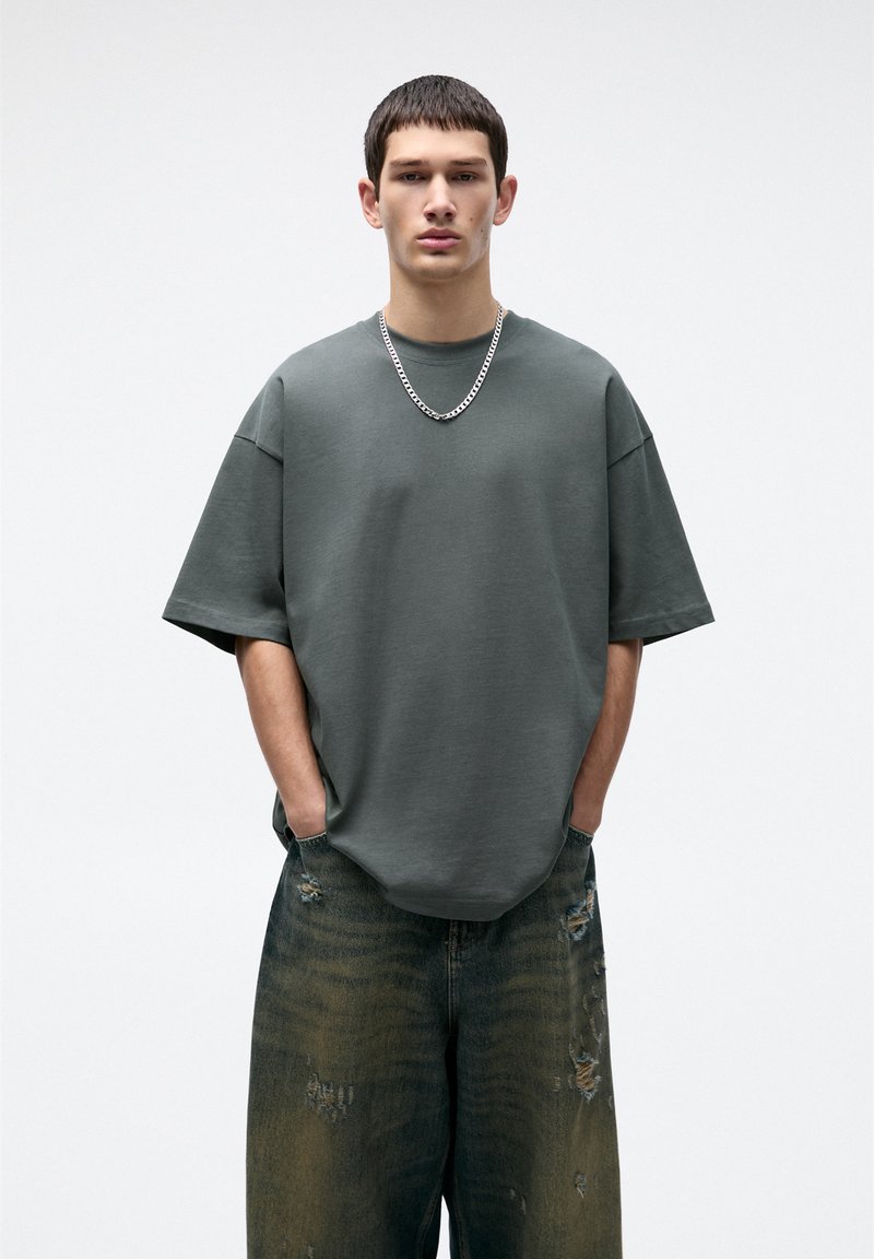 T-shirt oversize di un verde scuro in un tessuto morbido, abbinato a jeans a gamba larga con effetto distressed che presentano un gradiente dal scuro al chiaro.
