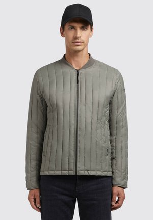 Quilted gray jacket with a round collar and front zipper, featuring vertical stitched detailing and side pockets. Worn with dark jeans and a black cap.