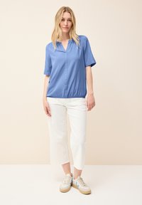 Blusa blu a maniche corte con scollatura a V e orlo arricciato, abbinata a pantaloni bianchi a taglio corto e sneakers beige con dettagli blu.