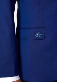 Zils bleizers ar gludu tekstūru, ar kabatu, kurā ir neliela izšūta rakstura dizains. Redzama balta aproce.