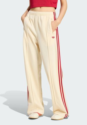 Blick von der Taille abwärts auf eine Person, die cremefarbene, weit geschnittene Trainingshose mit roten Seitenstreifen trägt, Hände in den Taschen, kombiniert mit weißen Schuhen und einem roten Crop-Top.