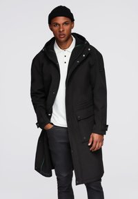 Abrigo largo negro con capucha, con botones a presión, bolsillos laterales y un tejido texturizado. Lleva puesto sobre una camisa tipo polo blanca y pantalones oscuros.