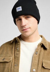 Zwarte gebreide beanie met een wit label patch bovenop een corduroy shirt in lichtbruin en een beige crewneck t-shirt eronder. Close-up portret.