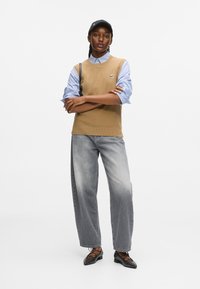 Kobieta stojąca z założonymi rękami, ubrana w czapkę w kolorze czarnym, beżowy sweter bez rękawów na niebieską koszulę w kratę, szare szerokie spodnie dżinsowe i czarne czółenka na spiczastej podeszwie.