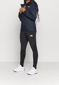 Laivastonsininen vedenpitävä takki hupulla, pitkät hihat; mustat vartalonmyötäiset legginsit väriblokeilla; valkoiset urheilukengät verkkokangasyksityiskohdilla.