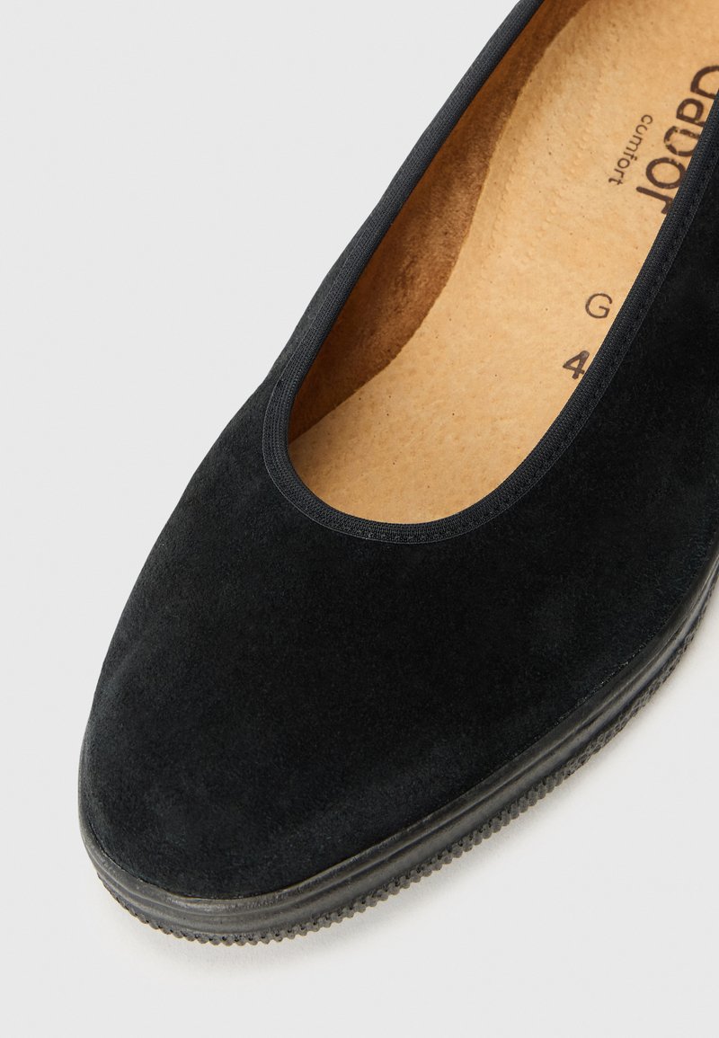 Ballerine noire en suède avec un bout rond, bord élastiqué et semelle en caoutchouc texturé. L'intérieur présente une semelle intérieure rembourrée beige.