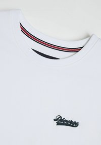 Biała bawełniana koszulka z okrągłym dekoltem, z czarnym haftowanym logo. We wnętrzu dekoltu znajduje się czerwono-czarny paskowany detal.