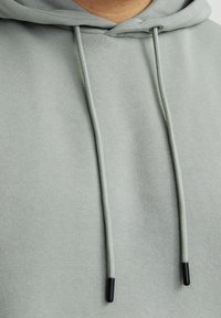 Lysegrøn hoodie med blød tekstur, snore i samme farve med sorte spidser og et enkelt, rent design.