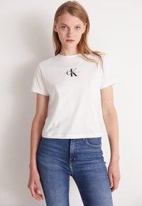 Balta медvilninė marškinė su trumpomis rankovėmis ir apvaliu kaklu. Priekinėje pusėje yra juodas Calvin Klein Jeans logotipas.