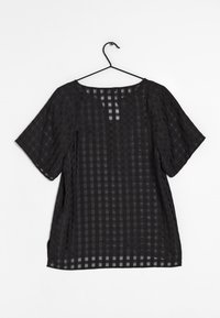 Blouse noire à manches courtes avec un motif transparent à carreaux, suspendue sur un cintre noir contre un fond blanc.