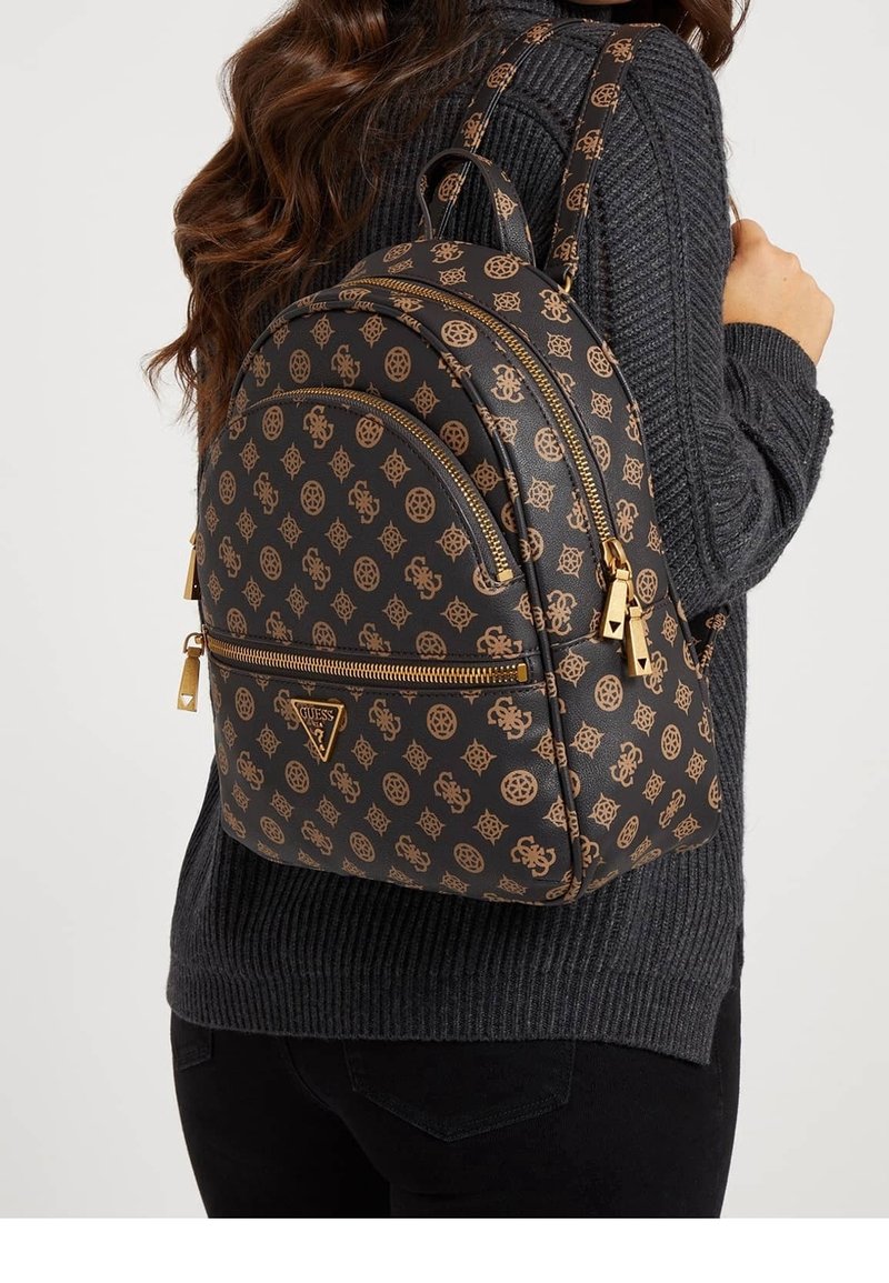 Femme portant un sac à dos Guess noir et marron à motifs avec des fermetures éclair dorées sur un pull en maille gris foncé et un pantalon noir.