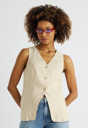 Göndör hajú nő piros napszemüvegben, bézs színű, gombos, ujjatlan mellényt visel és kék farmert, fehér háttér előtt áll.