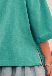 Kurzärmeliges türkisfarbenes Oberteil mit strukturierter Oberfläche, seitlichen Schlitzen und lockerem Schnitt, kombiniert mit einem hellen Jeansrock.