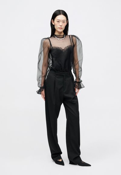 Μαύρη διαφανής μπλούζα με φουσκωτά μανίκια πάνω από ένα μονόχρωμο μαύρο τοπ, συνδυασμένη με ψηλόμεσα μαύρα παντελόνια και παπούτσια με μυτερή μύτη.