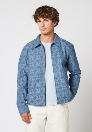 Homme portant une veste zippée bleue à motifs sur une chemise bleu clair et un pantalon blanc, debout devant un fond uni, les mains dans les poches.
