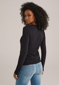 Kvinna med lockigt hår iförd en smalsvetsad svart långärmad tröja och ljusblå jeans, som tittar över sin axel mot en enkel bakgrund.